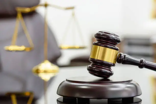 Photo of Wooden judge gavel or a wood hammer and a soundboard used by a judge person on a desk in a courtroom with a blurred brass scale of justice behind.