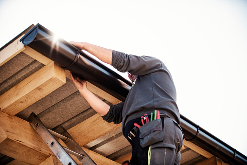 Roofer builder worker finishing folding gutter. He is up a ladder, photo taken from ground looking up, low angle view. He wears a tool belt, sky and clouds