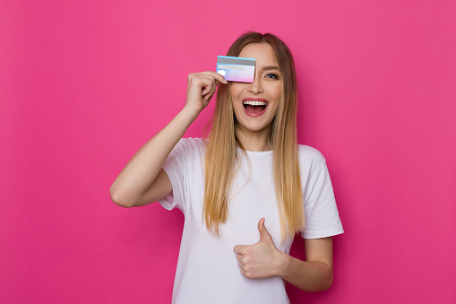 Laughing young woman in white shirt covers eye with credit card and gives thumb up. Waist up studio shot on pink background.