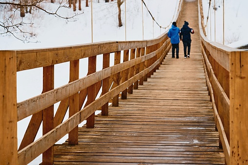 two teenagers walk together next to an old pedestrian suspended wooden bridge and light snow falls