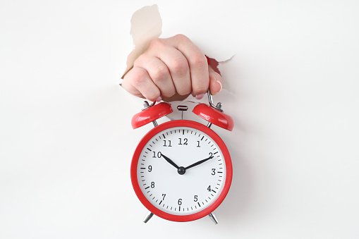 Red alarm clock holds hand through hole in white paper. Time management and business deadlines concept