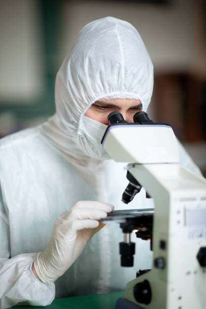 istock Scientist in coveralls and protective face mask and gloves looking through microscope binoculars 1310426492