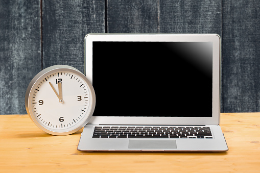 Laptop with blank screen, clock with arrows at five to twelve on a wooden table. mockup for your text