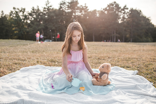 Beautiful blonde young girl playing with toys and teddy bear on picnic in a park on hot summer day