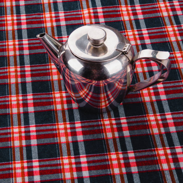 Stainless steel teapot on a table with a tablecloth with a pattern in a cage, top view.