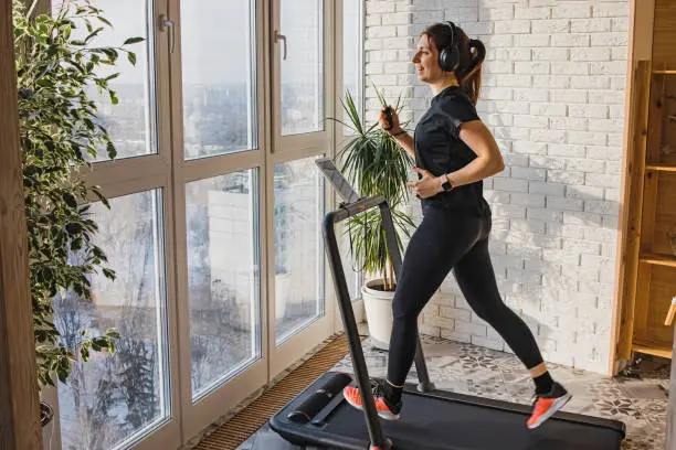 Young woman jogging on the modern compact treadmill at her home at room with big windows. Modern lifestyle, sport indoors.