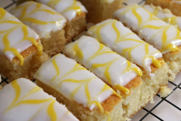 Stock photo showing close-up, elevated view of cooling rack of home-made lemon drizzle cake cut into slices. The lemon cake is topped with a white fondant icing and decorated with feathered, yellow, lemon flavoured icing. imagen de primer plano de la bandeja de limón casera hornear rebanadas de pastel cubiertas con glaseado de fondant blanco y glaseado con sabor a amarillo, limón, en el estante de refrigeración de metal, centrarse en primer plano, vista elevada