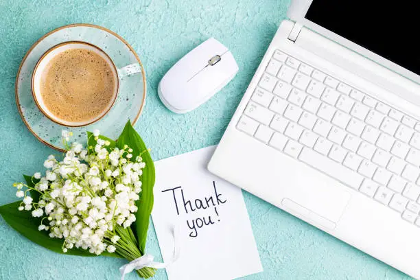 Photo of Notes thank you, coffee cup, laptop, bouquet of flowers lily of the valley on desk. Workplace in home office laptop and thank you note. Thankfulness, customer service, thanks card