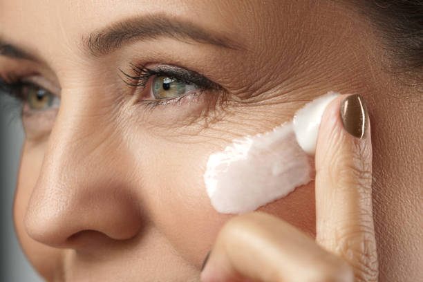 middle aged woman applying anti-aging cream on her face - wrinkled skin imagens e fotografias de stock