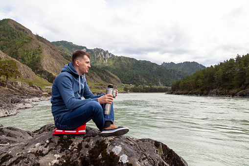 A brunette man of Caucasian appearance sits on a stone near the Katun river in the Altai mountains and drinks tea pouring into a mug from a thermos on a cool autumn day.