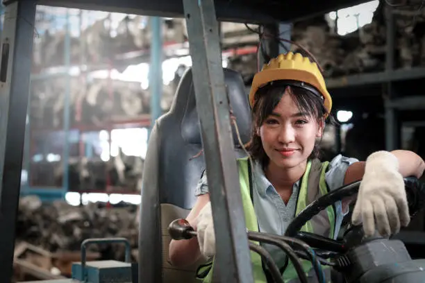 Industrial worker woman wearing helmet driving forklift car at manufacturing plant factory industry, Asian beautiful female engineer and many engine parts as blurred background