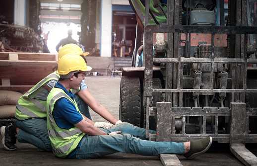 Accident at workplace, industrial engineer worker wearing helmet hit by forklift car at manufacturing plant factory construction site building, man leg stuck in forklift, colleague try to help him