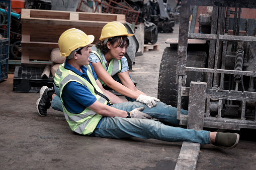 Accident at workplace, industrial engineer worker wearing helmet hit by forklift car at manufacturing plant factory construction site building, man leg stuck in forklift, colleague try to help him