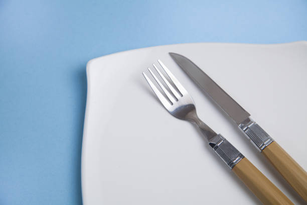 istock Clean empty white plate with knife and fork, on an isolated blue background. 1310478371