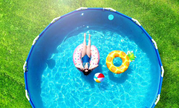 Aerial. Girl resting in a metal frame pool with inflatable toys. Summer leisure and fun concept. Frame pool stand on a green grass lawn. Top view from drone. stock photo
