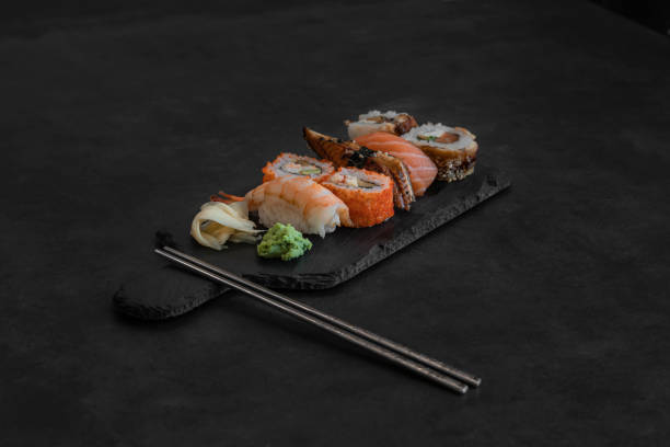 fermez-vous vers le haut du sushi de sashimi et du rouleau de sushi réglé avec des baguettes sur le plateau noir sur la photo foncée de stock de fond - sushi styles photos et images de collection