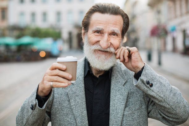 istock Mature man touching grey beard and holding coffee outdoors 1310485262