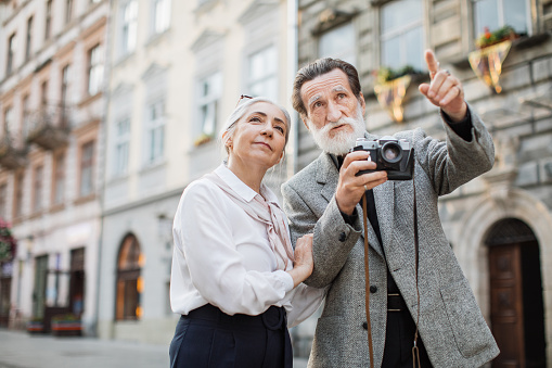 Beautiful mature bearded man with retro photo camera shows his wife architectural monuments. Senior family enjoying travelling time during retirement. Spending time together.