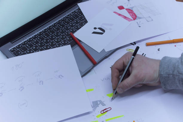 istock Creative workplace of a Graphic Designer. A man in the office is developing a logo on the table against the background of printed sketches and a laptop. 1310490203