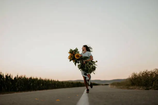 Female in a blue dress running in the middle of a countryside road and holding a bunch of fresh yellow flowers.