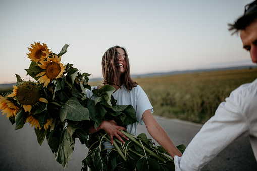 Beautiful spontaneous photo of a young woman in a blue dress, carrying sunflowers and holding hands with her handsome boyfriend. Enjyoing a walk on a sunny day down the countryside road, both smiling