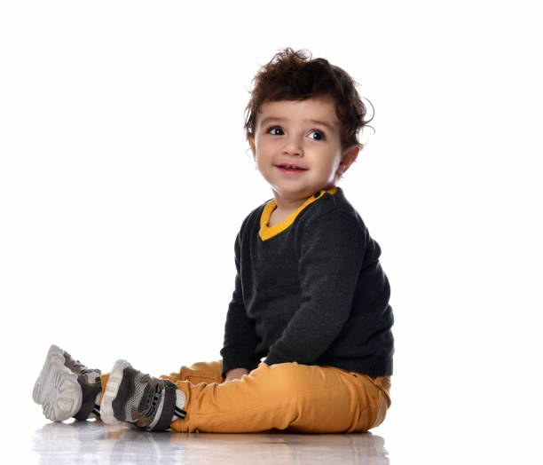 Infant baby boy toddler in white bodysuit is sitting sideways on the floor looking up at us on a gray background with free text copy space.