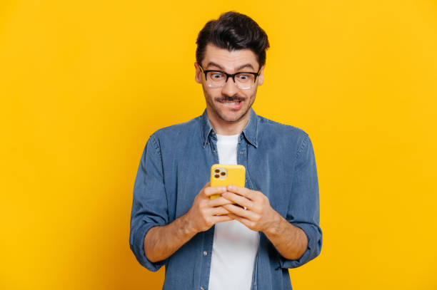 istock Happy handsome excited stylish guy using smartphone, chatting online, writing message, browsing internet, social media, looks surprised at cellphone, standing on isolated orange background 1310500776