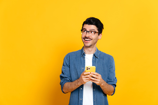 Joyful happy good looking stylish caucasian unshaven guy holding smartphone in hand, chatting online, browsing internet, looking happily to the side, standing against isolated orange background, smile