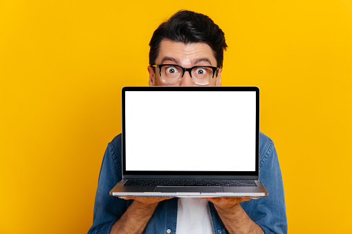 Amazed excited caucasian guy with glasses peeking out from behind laptop, looks surprised at camera, stands on isolated orange background, holds an open laptop with blank white screen, copy space