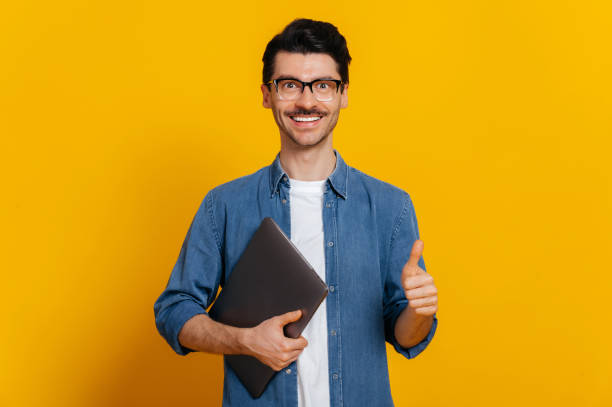 istock Happy confident caucasian stylish handsome guy with glasses, freelancer or student, holds a laptop in hand, shows a thumb up gesture, stands on isolated orange background, looks at camera, smiling 1310500827