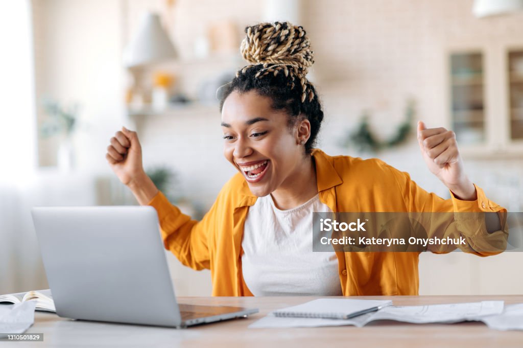 Overjoyed excited african american girl with dreadlocks, freelancer, manager working remotely at home using laptop, looks at screen with surprise, smiling face, gesturing with hands, got a dream job Computer Stock Photo