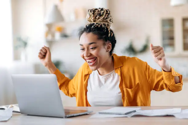 Photo of Overjoyed excited african american girl with dreadlocks, freelancer, manager working remotely at home using laptop, looks at screen with surprise, smiling face, gesturing with hands, got a dream job