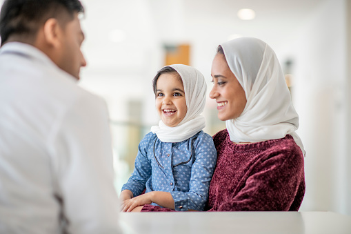 An adorable little girl wearing a hijab is sitting on her mother's lap at a medical check up appointment with their family doctor. She is smiling at the doctor who is sitting across the table.