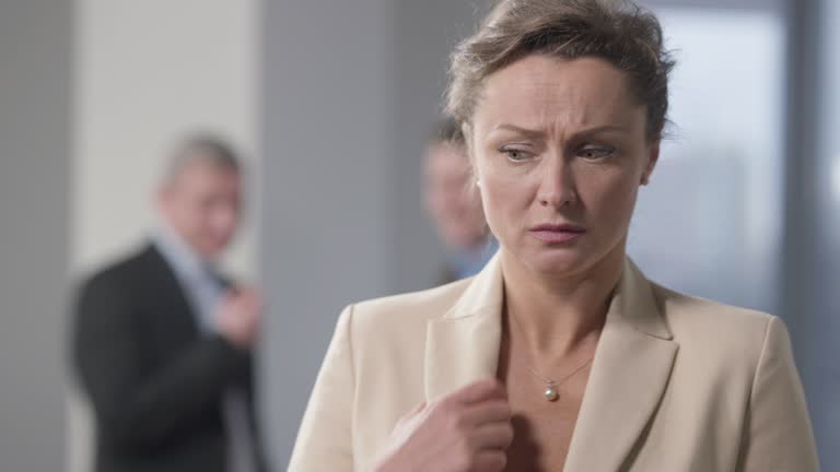 Stressed brunette woman with hazel eyes standing indoors thinking with blurred men talking at background. Portrait of bullied Caucasian female employee in office. Discrimination and harassment.