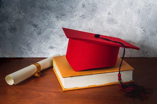 Still life with a red graduation hat on a book next to a diploma on a wooden desk. Concept of studies and graduation