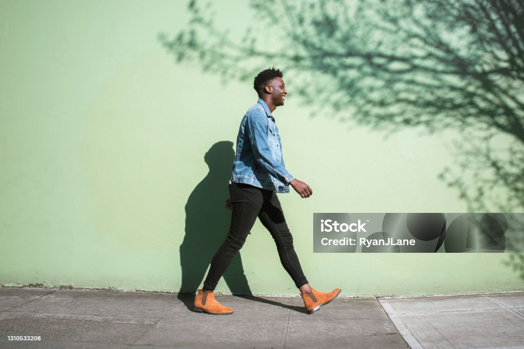 Happy Adult Man Walking Sunny City Street Sidewalk A young adult African American enjoys the sunshine as he strolls down a city walkway.  The bright sunlight casts interesting shadows on the wall behind. Walking Stock Photo