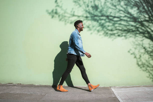 Happy Adult Man Walking Sunny City Street Sidewalk A young adult African American enjoys the sunshine as he strolls down a city walkway.  The bright sunlight casts interesting shadows on the wall behind. walking stock pictures, royalty-free photos & images