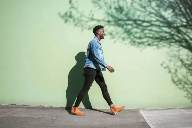 A young adult African American enjoys the sunshine as he strolls down a city walkway.  The bright sunlight casts interesting shadows on the wall behind.