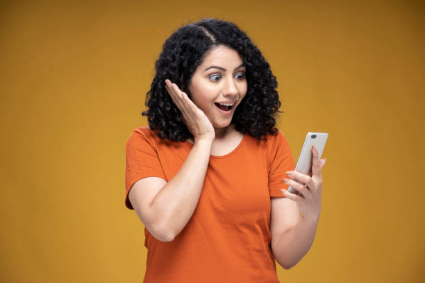 shot of a young women using  mobile phone social media, surfing the net standing isolated over yellow background:- stock photo Adult, adult only,  social media, surprise, shock, surfing the net, using phone, India, Indian ethnicity, cheek cell stock pictures, royalty-free photos & images