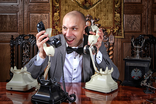 Fast-paced desperate businessman showman works in office and many calls, he keeps lot handsets. Business management concept. Stressed man talks on many phones at once. Space for inscription or logo