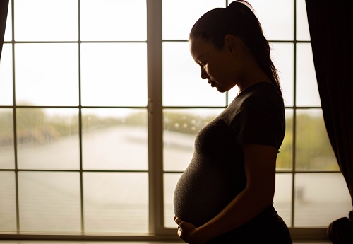 A sad pregnant woman standing near a window looking down depressed.