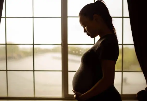 A sad pregnant woman standing near a window looking down depressed.