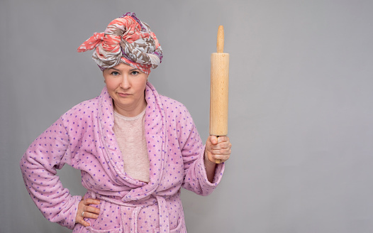 A woman in a dressing gown and a scarf on her head holds a rolling pin in her hand and looks seriously into the camera. Woman 30-40 years old in home clothes on a gray background, photo in the studio.