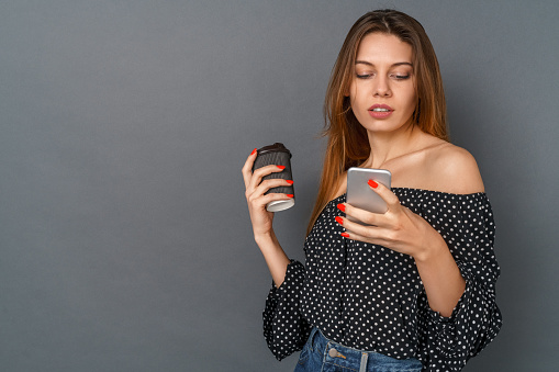 Young woman casual style studio isolated on gray wall browsing smartphone social media concentrated drinking hot coffee