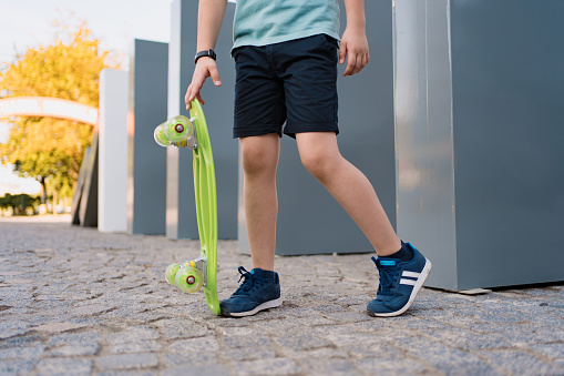 Close up legs in blue sneakers with green skateboard. Active urban lifestyle of youth, training, hobby, activity. Active outdoor sport for kids