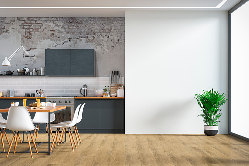 Empty anthracite modern kitchen  on hardwood floor with appliances, full dining table, chairs in front of white ruined brick wall background. A potted plant in front of a large white plaster wall background with copy space and windows on a side. 3D rendered image.