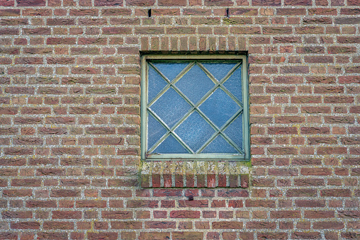 Old square window with glazing bars in a masonry brick wall. The light green paint has peeled off. The photo was taken at an old factory building in the Netherlands.