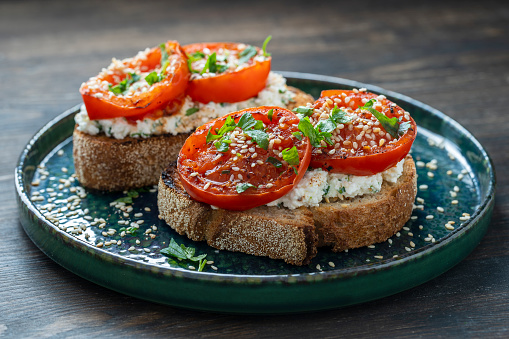 Delicious toasted bread with roasted red tomatoes, feta cheese, sesame seeds and herbs on plate, close up
