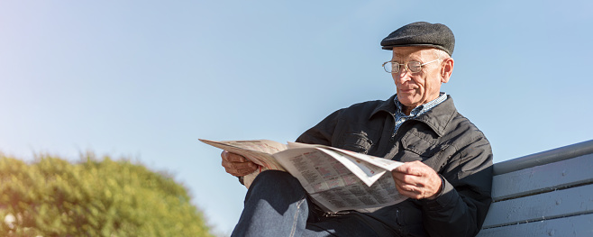 Adult citizen in cap and eyewear seated on bench and reads newspaper in urban park outdoor.