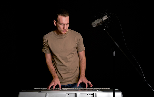 Young caucasian man sing while playing an electric keyboard in front of black soundproofing walls. Musician producing music in professional recording studio.